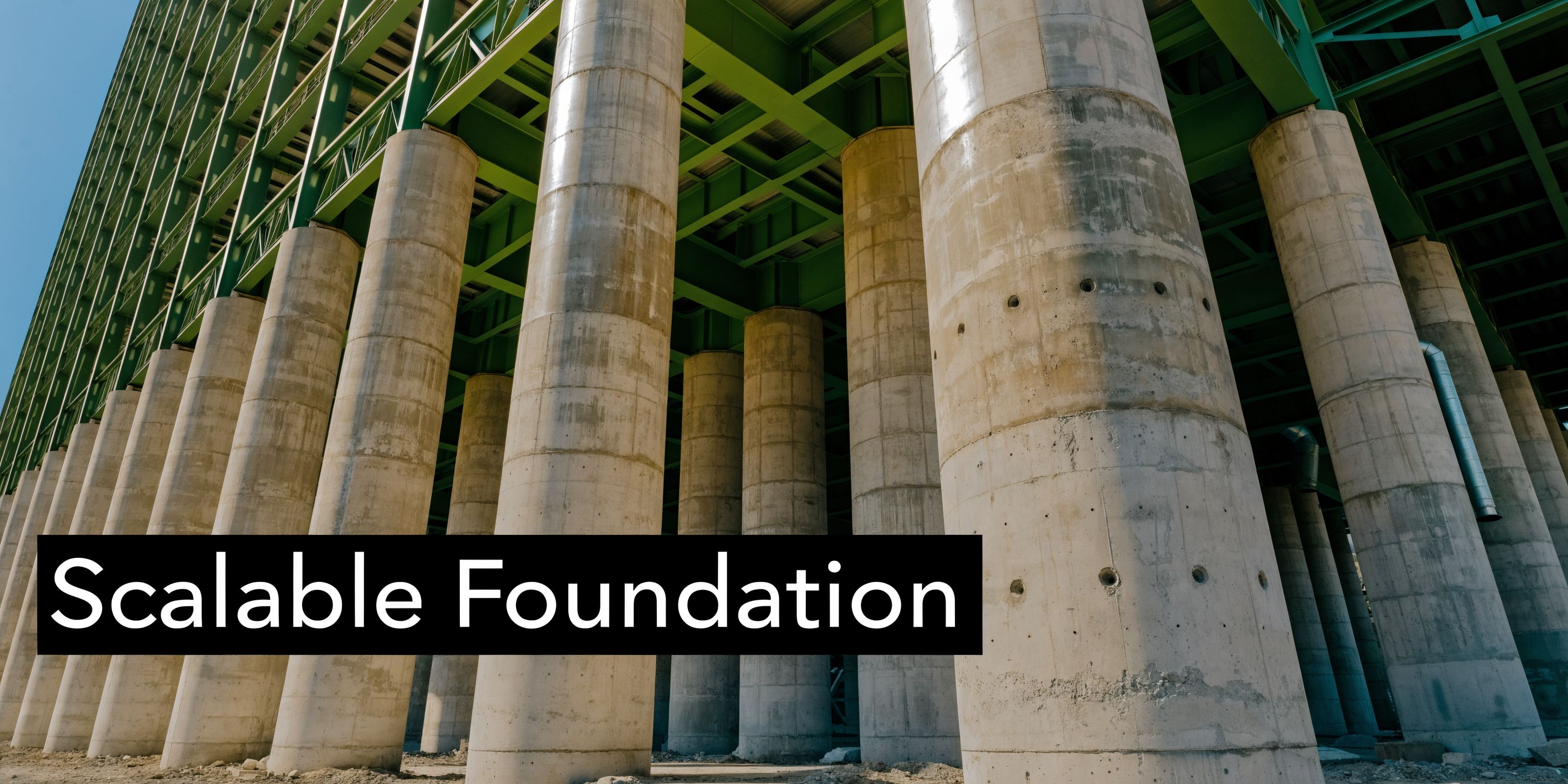 A low-angle view of massive concrete bridge pillars supporting a large green steel industrial structure overhead.